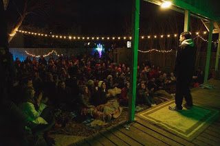 A man dressed in black with a white hoodie stands on an elevated porch serving as a stage illuminated in green light and tells his story to a crowd of about 100 people seated cozily on blankets on the ground in a backyard. Audience members at left clap. Fiesta style lights hang in the air over the audience