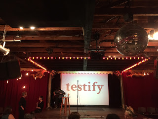 Two female sign language interpreters dressed in black sign to each other. To the right of the interpreters is an elevated stage on which a man plays acoustic guitar. Also on stage is an empty stool and a microphone stand. Behind the stage the word "testify" in black text is projected over a white screen. Red curtains cover the walls on either side of the stage. A disco ball hangs in the foreground